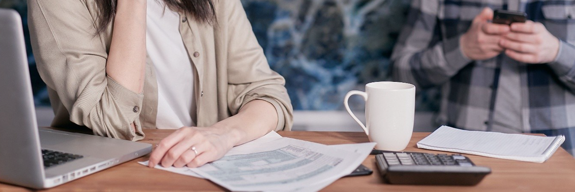 Work desk scene: a person sits with a laptop and papers while another person uses a phone; mug, notebook and calculator on the table.
