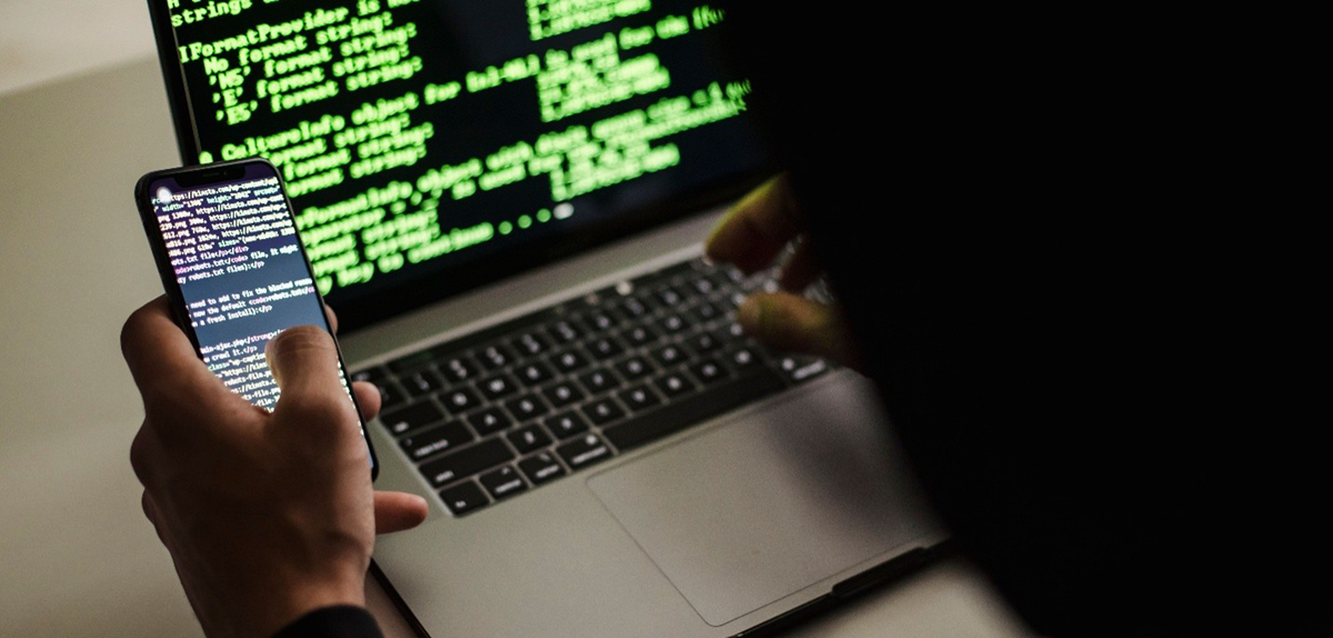 Person holding a smartphone displaying code while typing on a laptop with green terminal text on screen (coding scene).